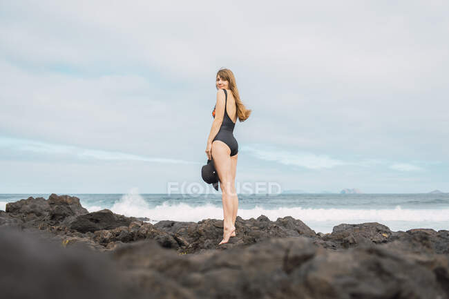 З боку погляд привабливої жінки в купальнику, яка тримає капелюх і дивиться на камеру, стоячи на кам'яному березі біля махаючи морем проти переповненого неба. — стокове фото