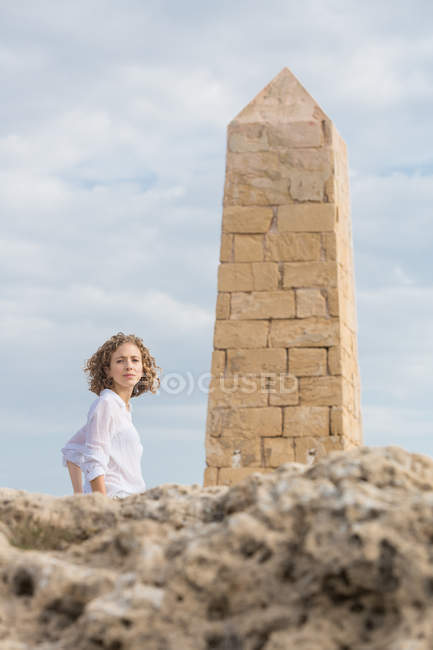 Jeune femme coûteuse regardant la caméra près de la construction en pierre en forme de tour sur la roche — Photo de stock