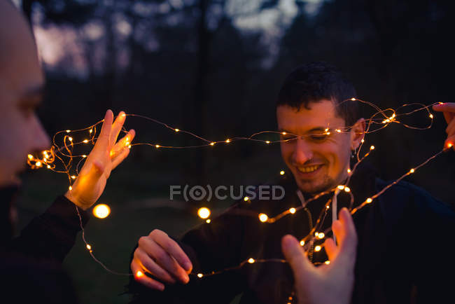 Closeup of men untangling illuminated fairy lights in murk forest in evening on blurred background — Stock Photo