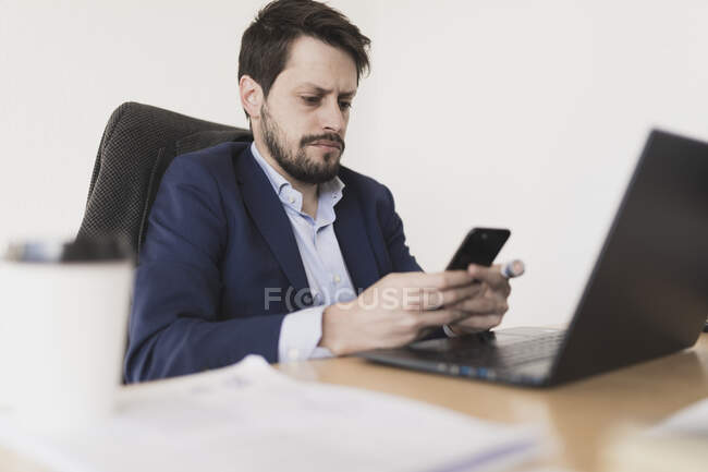 Концентрований молодий чоловік розмовляє на мобільному телефоні і переглядає ноутбук за столом в офісі — стокове фото