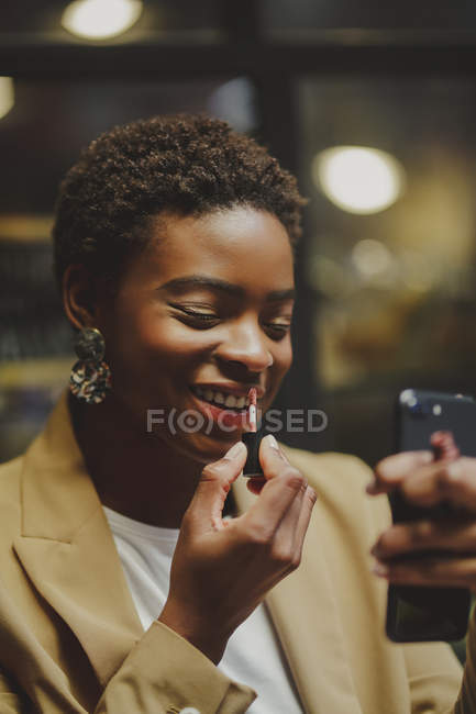 Mujer elegante afroamericana poniendo lápiz labial en los labios y sosteniendo el teléfono móvil en la calle - foto de stock