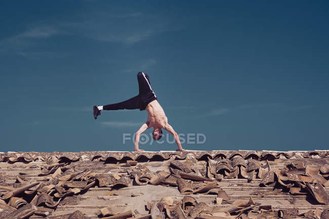 Junger Mann ohne Hemd beim Handstand auf verwittertem Dach eines alten Gebäudes vor blauem Himmel — Stockfoto