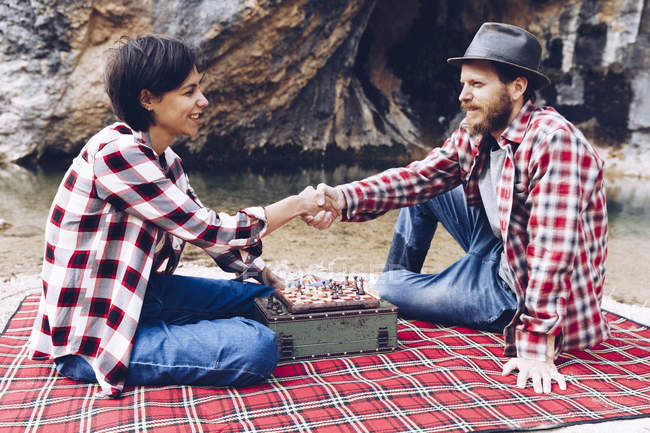 Side view of man and woman in plaid shirts playing chess on plaid having picnic on shore of lake in cliffs — Stock Photo