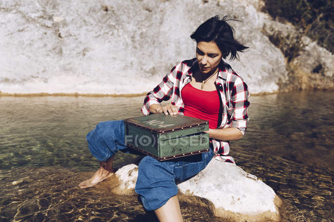 Mujer sentada en la roca en el agua transparente del lago mirando dentro de la vieja caja de metal oxidado teniendo picnic - foto de stock