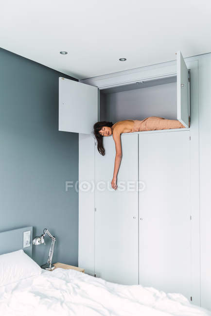 Young sensual woman sleeping on high shelf of wardrobe near bed in bedroom — Stock Photo