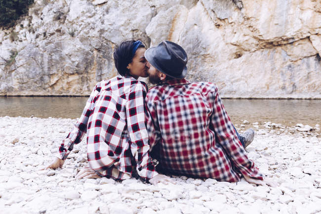Vista trasera de pareja joven sentada y besándose en la costa rocosa del río de montaña cerca del acantilado - foto de stock