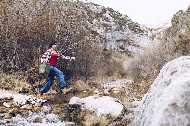 Vue latérale d'une femme occasionnelle portant étui et sautant sur des rochers de ruisseau clair de l'eau dans la nature — Photo de stock