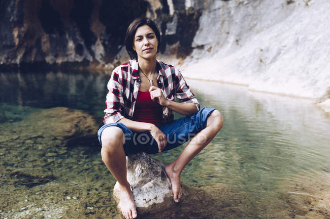 Feliz mujer adulta sentada en la roca en el tranquilo agua transparente del lago disfrutando de la naturaleza y sonriendo - foto de stock