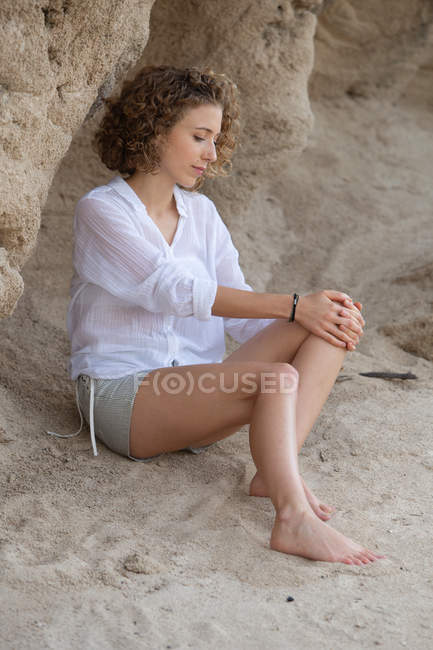 Joven mujer de ensueño en camisa blanca y pantalones cortos sentados en la playa cerca de la roca - foto de stock
