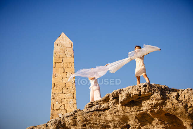 Desde abajo mujeres misteriosas jóvenes con las manos levantadas sosteniendo textiles blancos y posando sobre rocas y cielo azul - foto de stock