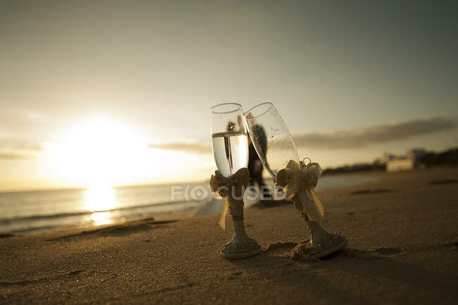 Copos de close-up de champanhe na costa de areia e recém-casados abraçando perto da água ao pôr do sol no fundo borrado — Fotografia de Stock