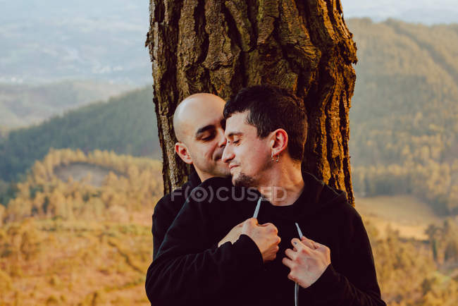Alegre casal homossexual abraçando perto da árvore na floresta e vista pitoresca do vale — Fotografia de Stock