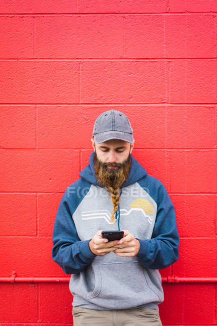 Attractive guy with braided beard browsing smartphone while leaning red wall on city street — Stock Photo