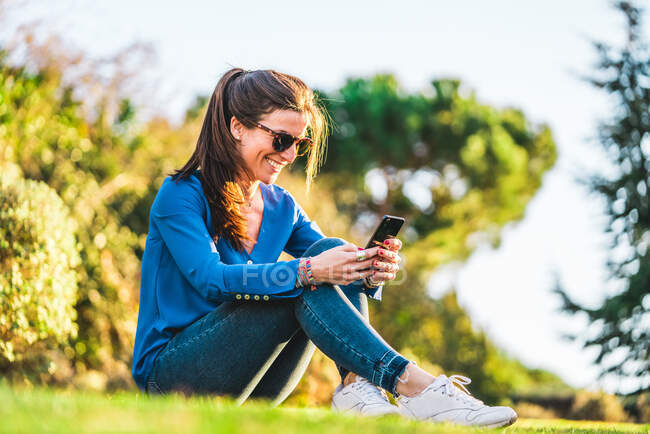 Portrait de jeune femme avec des lunettes de soleil en utilisant smartphone assis — Photo de stock