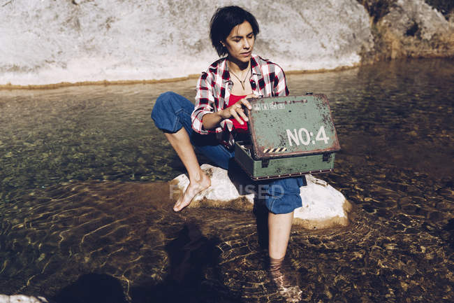 Femme assise sur la roche dans l'eau transparente du lac regardant à l'intérieur de vieux boîtier en métal rouillé ayant pique-nique — Photo de stock