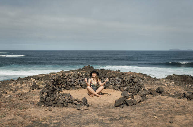 Mujer con estilo en traje de baño y sombrero sentado dentro del círculo de piedra y meditando mientras pasa tiempo cerca de ondear el mar en el día nublado - foto de stock