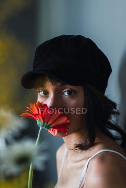Jovem mulher morena encantadora em boné olhando para a câmera e segurando flor vermelha fresca no quarto — Fotografia de Stock
