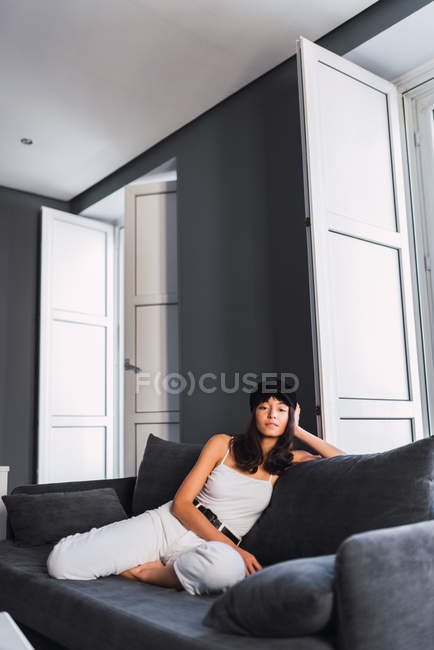 Приваблива молода стильна жінка в Кап дивлячись на камеру, відпочиваючи на дивані біля балкона будинку — стокове фото