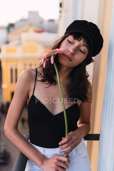 Молоді тонкий жінка в Кап з свіжою квіткою дивиться геть, стоячи на балконі на розмитий фон — стокове фото