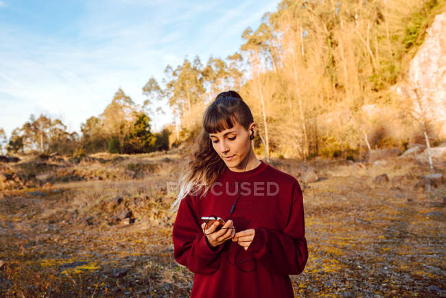Mulher jovem hipster com piercing e fones de ouvido ouvindo música com telefone celular e andando na estrada rural — Fotografia de Stock