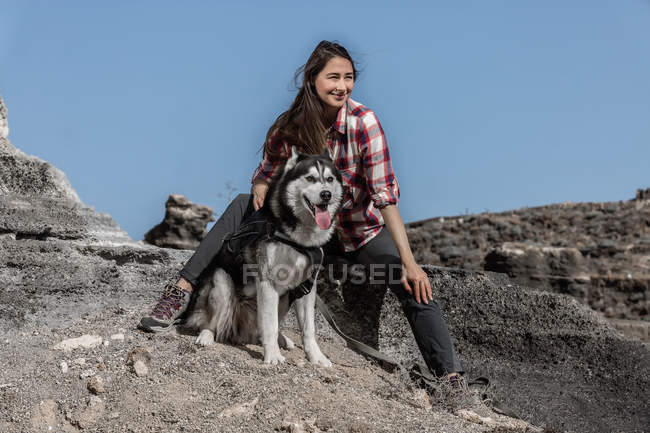 Cheerful young woman sitting near husky on rock in nature — Stock Photo