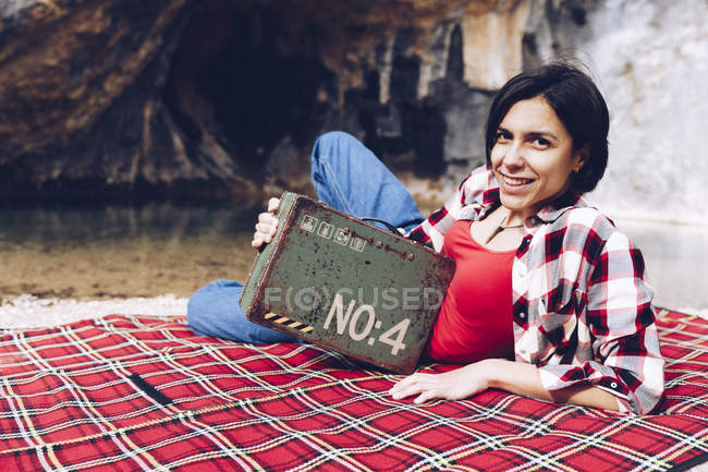 Mujer adulta sonriente acostada sobre cuadros en la orilla del lago en acantilados que sostienen un viejo caso oxidado con provisión sonriendo a la cámara — Stock Photo