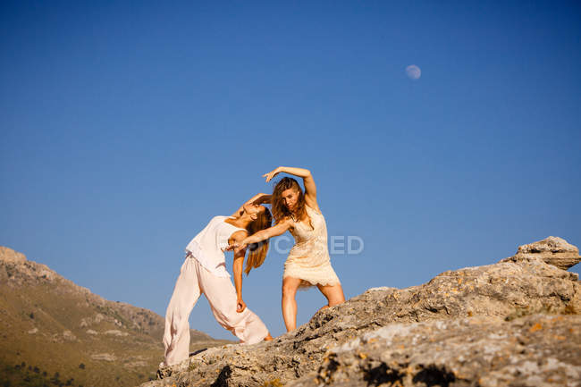 Молодые загадочные женщины с поднятыми руками позируют на скалах возле холма и голубого неба с луной — стоковое фото