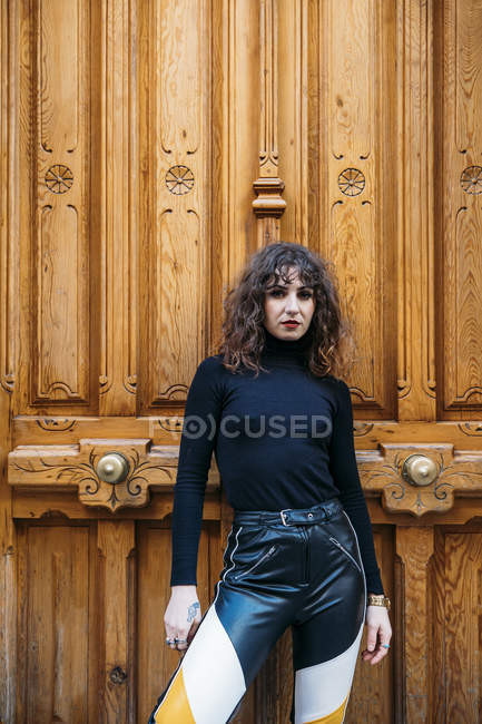 Donna in abito elegante guardando la fotocamera mentre in piedi vicino alla porta in legno ornamentale di vecchio edificio sulla strada della città — Foto stock