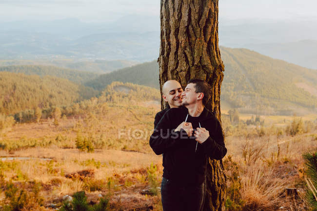 Веселая гомосексуальная пара, обнимающая дерево в лесу и живописный вид на долину — стоковое фото