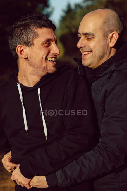 Retrato de alegre casal homossexual abraçando na floresta em dia ensolarado no fundo borrado — Fotografia de Stock