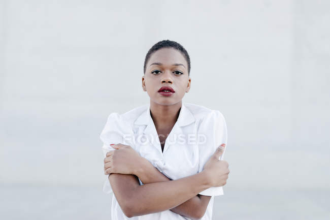 Moda modello etnico a pelo corto in camicia bianca con le braccia incrociate in posa contro muro grigio — Foto stock