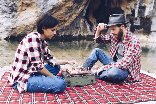 Seitenansicht von Mann und Frau in karierten Hemden beim Schach spielen auf karierten Picknick am Ufer des Sees in den Klippen — Stockfoto