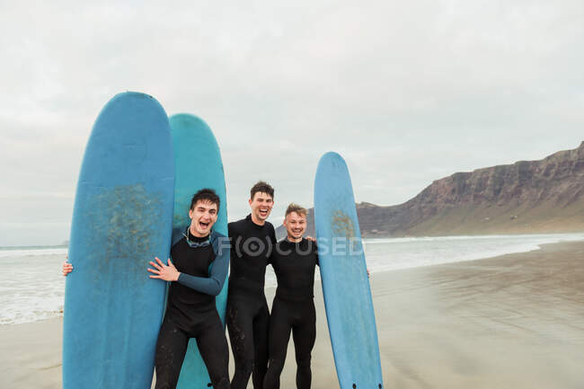 Три молоді чоловіки в чорному купальнику сміються і дивляться на камеру, тримаючи блакитні дошки для серфінгу і стоячи на березі біля моря — стокове фото