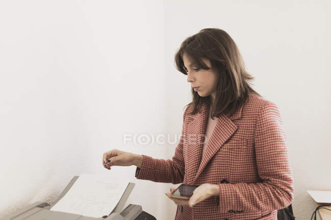 Jeune femme concentrée tenant un téléphone portable et imprimant des documents au bureau — Photo de stock