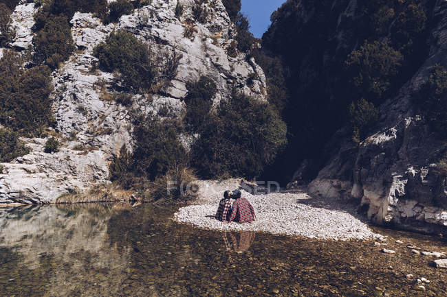 Vista trasera de pareja joven sentada y besándose en la costa rocosa del río de montaña cerca del acantilado - foto de stock