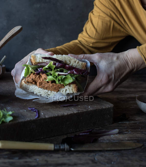 Mãos de colheita de pessoa que mantém o sanduíche de patê de tomates, salada fresca e repolho na bandeja perto de faca na tábua de madeira — Fotografia de Stock
