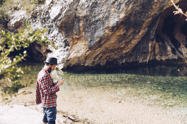 Side view of traveling man using phone while standing on sandy shore of tranquil lake against cliff — Stock Photo