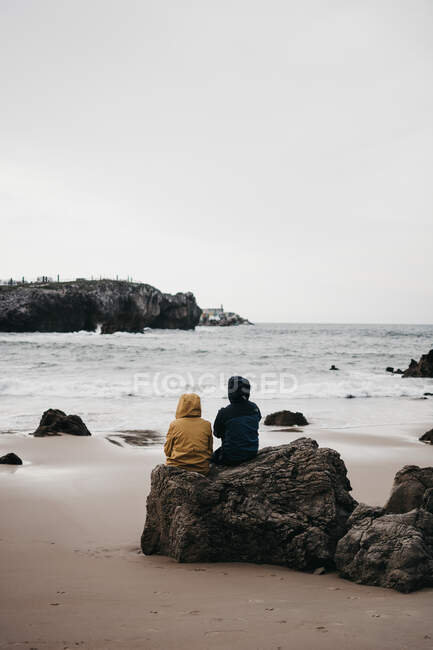 Vista trasera de dos personas en chaquetas calientes sentadas en rocas ásperas y mirando juntos el mar tormentoso - foto de stock
