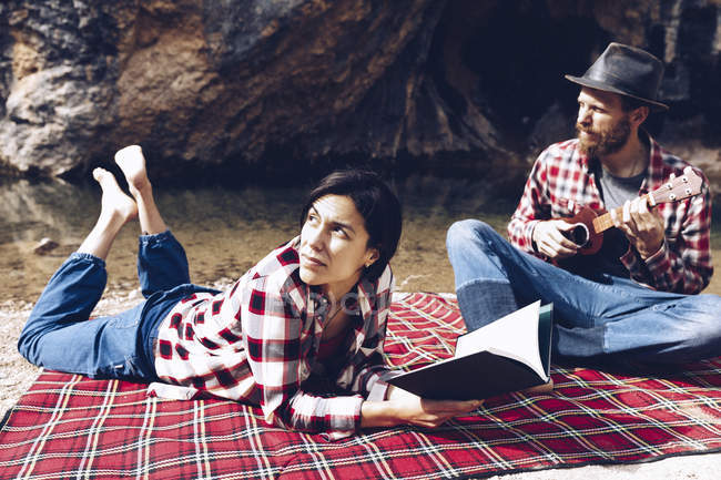 Adult man and woman on checkered plaid with book and small ukulele having picnic on lake shore — Stock Photo