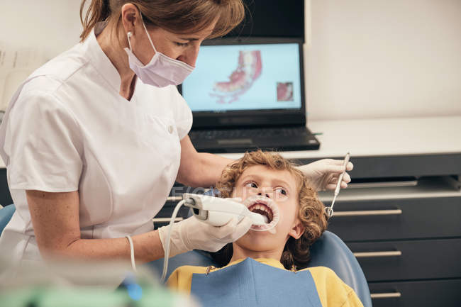 Femme en masque et uniforme de médecin faisant le balayage des dents du petit garçon tout en travaillant dans la clinique dentaire — Photo de stock
