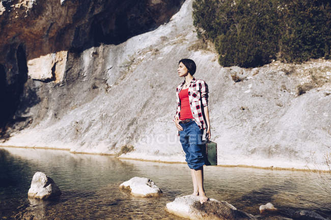Женщина, стоящая на скале с футляром рядом с чистой водой в озере — стоковое фото