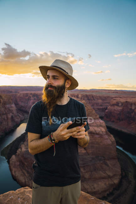 Человек в шляпе на мобильном телефоне, стоя на фоне каньона и реки во время заката на Западном побережье США — стоковое фото