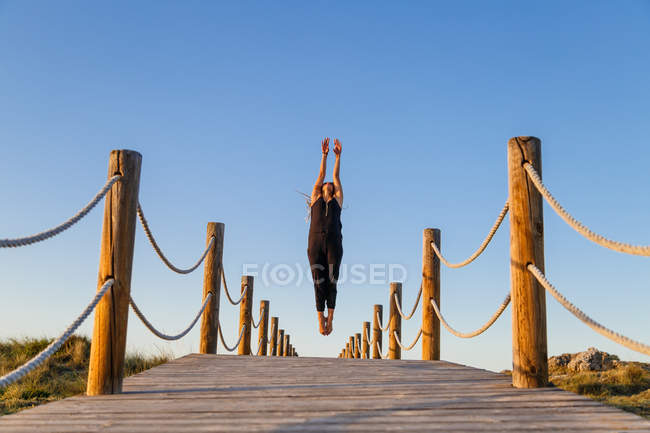Junge Ballerina in schwarzer Kleidung mit erhobenen Armen in der Luft auf einem Steg und blauem Himmel bei sonnigem Tag — Stockfoto