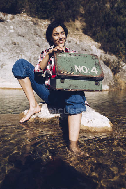 Женщина, сидящая на камне в прозрачной озерной воде, глядя внутрь ржавого металлического корпуса, устраивает пикник — стоковое фото