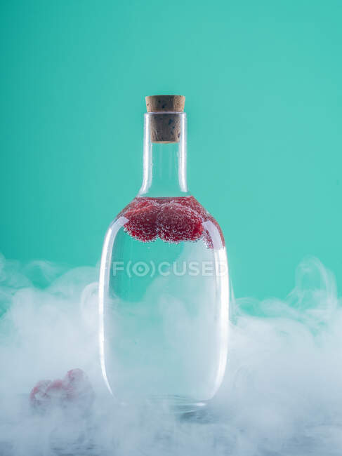 Garrafa de vidro com bagas e álcool a bordo entre a névoa no fundo azul — Fotografia de Stock