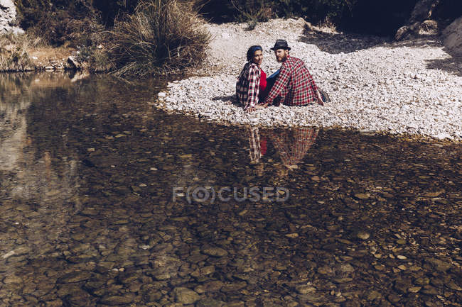 Вид збоку молодої пари, що сидить на скельному узбережжі гірської річки біля скелі — стокове фото