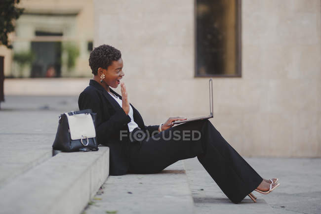 Улыбающаяся уверенная афро-американская элегантная женщина в костюмах, просматривающая ноутбук и сидящая на лестнице рядом с сумкой на улице — стоковое фото