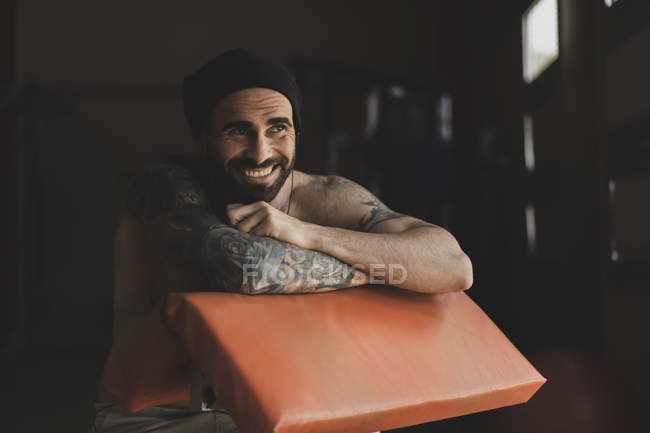 Красивий бородатий спортсмен з голим торсом посміхається і дивиться геть, сидячи на розмитому тлі тренажерного залу під час тренування — стокове фото