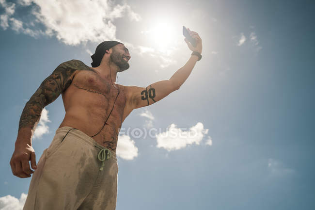 Fröhlicher muskulöser Sportler mit Smartphone gegen blauen Himmel mit Wolken — Stockfoto