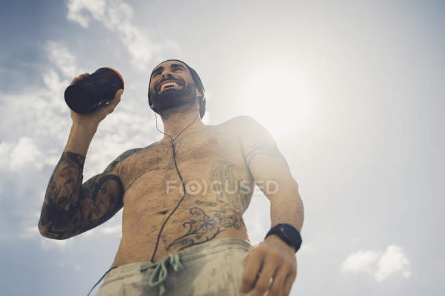 Uomo muscoloso senza maglietta che beve durante l'allenamento contro il cielo blu con le nuvole — Foto stock